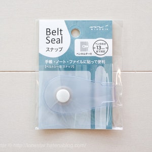 May include: A clear plastic belt seal snap for notebooks and journals. The snap is designed to hold up to 13 mm thick notebooks. The packaging shows the snap attached to a notebook.