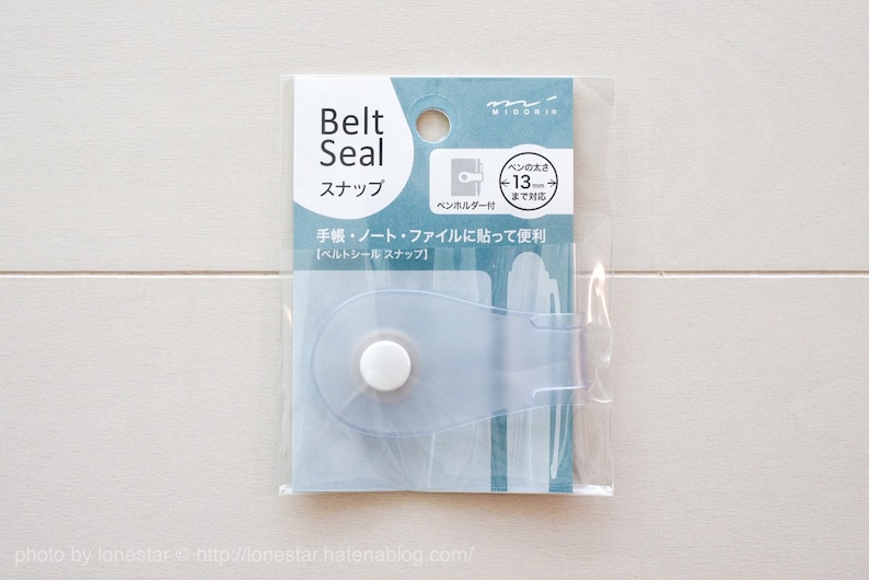 May include: A clear plastic belt seal snap for notebooks and journals. The snap is designed to hold up to 13 mm thick notebooks. The packaging shows the snap attached to a notebook.