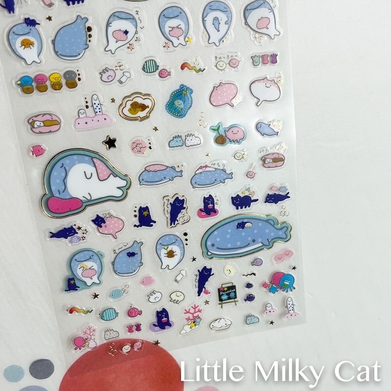 Puede incluir: Una hoja de pegatinas transparentes con varias ilustraciones de criaturas marinas y gatos en azul, rosa y dorado. Los dise&ntilde;os incluyen ballenas, gatos, peces y otra vida marina. El texto "Little Milky Cat" es visible en la parte inferior.