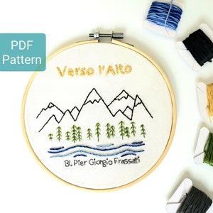 May include: A PDF pattern for an embroidery hoop with a mountain scene. The embroidery features black mountains, green trees, and blue water. The text "Verso l'Alto" is embroidered in gold thread. The text "Bl. Pier Giorgio Frassati" is embroidered in black thread.
