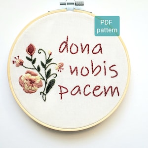 以下が含まれることがあります： 赤い糸で「Dona Nobis Pacem」という文字と花柄が刺繍されたフープアート。フープは木でできており、白い布地の背景があります。
