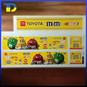 以下が含まれることがあります： トヨタ、M&M's、NASCARのロゴが入ったデカールシート。18番が大きく表示されています。M&M'sのキャラクターとスポンサーロゴのカラフルなグラフィックが特徴です。「TamiyaDecals.com」の文字が見えます。