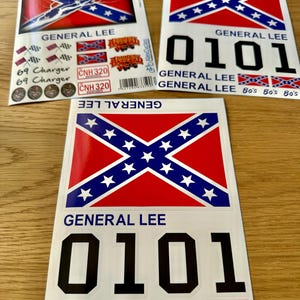 May include: Three stickers featuring the Confederate flag, the words "GENERAL LEE", and the number "0101". Additional text includes "69 Charger", "CNH 320", and "Bo's General Lee". The stickers are on a light brown surface.