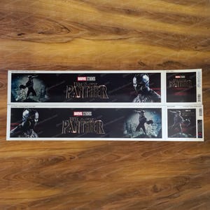 May include: Two Black Panther movie promotional stickers featuring the Marvel Studios logo and the Black Panther title in gold lettering. The stickers feature a black superhero character in a black suit with a black mask. The stickers are on a white background.