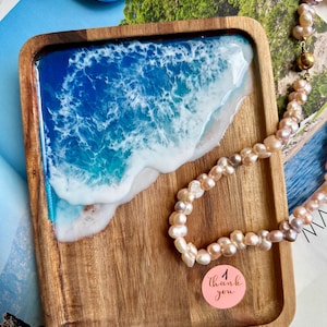 Könnte beinhalten: Ein rechteckiges Holztablett mit einer Harzeinlage, die Meereswellen in Blau-, Türkis- und Weißtönen nachahmt. Eine Kette aus hellrosa Perlen und ein runder rosa Aufkleber mit dem Text thank you liegen auf dem Tablett.