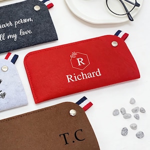 Puede incluir: Varios estuches personalizados para gafas en fieltro, en colores beige, gris, rojo y marrón. Cada estuche presenta texto o iniciales personalizados, con un detalle a rayas. Un par de gafas negras sobre un plato blanco.