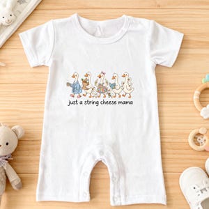 May include: White baby romper featuring a line of geese in outfits, with the text "just a string cheese mama". The romper has snap closures and short sleeves. The illustration depicts geese in various outfits.