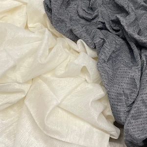 Può includere: Due pezzi di tessuto, uno bianco e uno nero, sono mostrati in primo piano. Il tessuto bianco è un materiale leggero e trasparente con un leggero luccichio. Il tessuto nero è un materiale più pesante e strutturato con un motivo intrecciato.