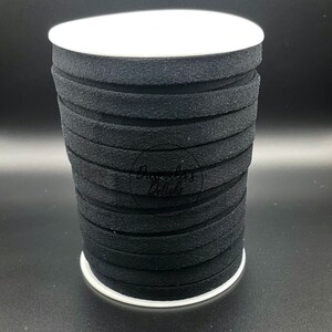 May include: A spool of black, flat, suede-like ribbon. The ribbon is stacked in a roll, with a white base and top. The ribbon is a solid black color and appears to be approximately 1/8 inch thick.