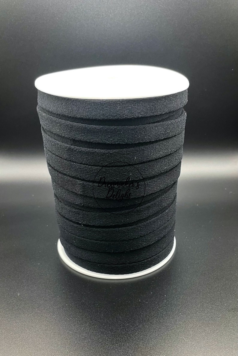 May include: A spool of black, flat, suede-like ribbon. The ribbon is stacked in a roll, with a white base and top. The ribbon is a solid black color and appears to be approximately 1/8 inch thick.