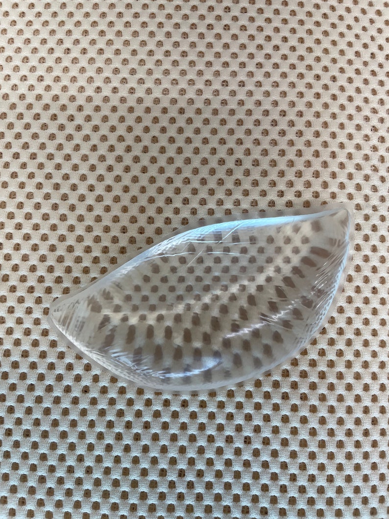 Puede incluir: Un objeto de pl&aacute;stico transparente y curvado, posiblemente una plantilla, est&aacute; envuelto en pl&aacute;stico transparente. El objeto se encuentra sobre una superficie de tela beige perforada, creando un contraste de texturas y colores.