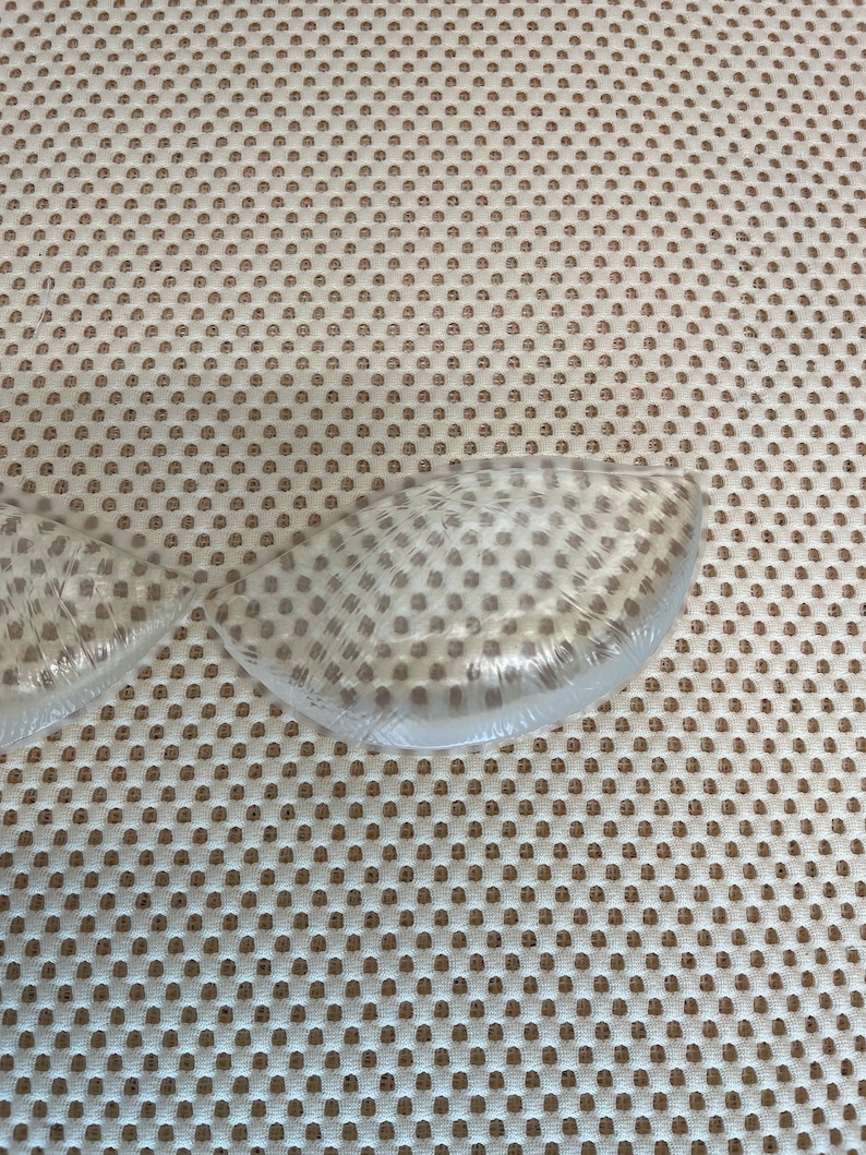 Puede incluir: Insertos de sujetador transparentes en forma de media luna en envase de pl&aacute;stico. Los insertos est&aacute;n hechos de un material suave y flexible. El fondo es una tela beige con un patr&oacute;n repetido de peque&ntilde;os agujeros espaciados.