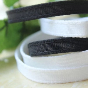 May include: Close-up of black and white ribbon. The black ribbon has a textured, ribbed appearance, while the white ribbon is smooth. The ribbons are layered, creating a visually appealing contrast. Ideal for crafting and sewing projects.