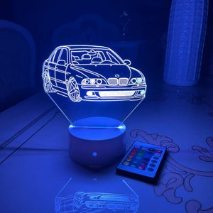 Könnte beinhalten: Eine weiße, 3D-gedruckte Nachtlampe in Form eines BMW-Autos. Die Nachtlampe wird mit blauem Licht beleuchtet und steht auf einem weißen Sockel. Eine Fernbedienung mit mehreren farbigen Knöpfen befindet sich im Vordergrund.