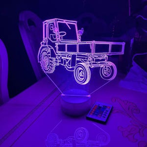 Peut inclure: Veilleuse LED en forme de tracteur. La lumière est un contour détaillé d'un tracteur avec une benne, éclairé en blanc froid. La lumière repose sur une base en bois et une télécommande est incluse.