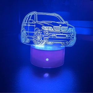 Op de afbeelding: Een neon nachtlampje in de vorm van een auto. De auto is een gedetailleerde omtrek, verlicht in helder wit tegen een donkerblauwe achtergrond. Het licht staat op een roze voet, wat een opvallend contrast creëert.