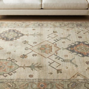 May include: Beige area rug with a geometric pattern in shades of brown, green, and orange. The rug features a central medallion design and a border with similar colors. The rug is placed on a hardwood floor.