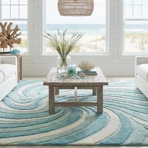 May include: A coastal-themed living room with white sofas, a wooden coffee table, and a blue and white wave-patterned rug. The room is bright and airy, with large windows overlooking a beach scene. Decorative items include coral and driftwood.