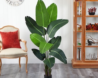 May include: A large artificial green plant with long, wide leaves in a black pot. The plant is in a room with a white wall and a wooden cabinet in the background.