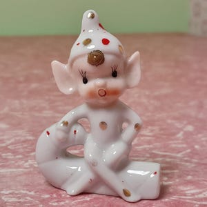 May include: A small, white ceramic elf figurine with large ears and a pointed hat, decorated with red and gold polka dots. The elf is seated on a white base with gold accents. A charming collectible, likely vintage.