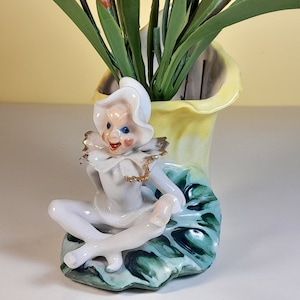 May include: A whimsical ceramic figurine of a jester with a white hat and blue eyes sits on a green and yellow leaf-shaped vase. The vase is filled with pink flowers.