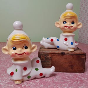 Puede incluir: Dos figuras de payaso de cerámica vintage. Los payasos son blancos con lunares rojos, azules y verdes. Tienen el pelo amarillo, mejillas rosadas y llevan sombreros blancos con una bola blanca encima. Un payaso está sobre una caja de madera.