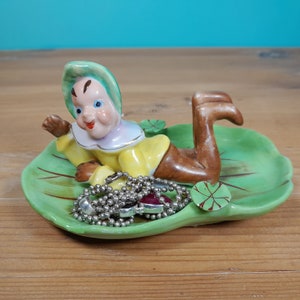 May include: A ceramic trinket dish with a reclining gnome figure on a green lily pad. The gnome is dressed in yellow, brown boots, and a green hat. A silver beaded necklace with a red gem is in the dish. The dish is a decorative item.