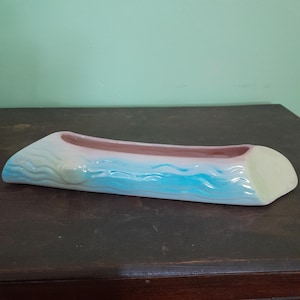 Puede incluir: Una jardinera de cerámica con forma de tronco, pintada con tonos azules, blancos y rosas. La jardinera tiene forma rectangular con extremos redondeados y una abertura en forma de canal. El diseño evoca un tema costero o náutico.