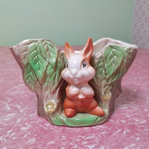 May include: A ceramic planter featuring a seated bunny figure. The bunny has white fur, orange ears, and a matching body. The planter is shaped like a tree trunk with green leaves and a neutral color palette.