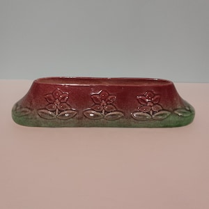 Puede incluir: Un macetero de cerámica rojo y verde con un diseño floral. El macetero tiene forma de rectángulo largo y estrecho.