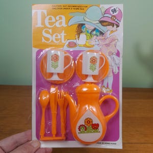 May include: A vintage orange plastic children's tea set, including a teapot, two cups and saucers, and cutlery. The packaging features illustrations of children and the text "Tea Set". The set is marked "Made in Hong Kong".