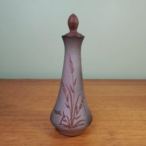 May include: A tall, slender decorative bottle with a brown wooden stopper. The bottle is a muted lavender colour with a brown wheat stalk design. The bottle is approximately 25 cm tall.