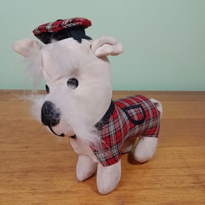 May include: A soft toy dog with a white body, black eyes and nose, and a red and black tartan outfit. The dog wears a matching tartan hat. The toy has a black collar and white fur around the face.