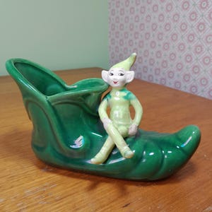 May include: A vintage ceramic planter shaped like a green shoe, with a seated elf figurine. The elf is dressed in green with a pointed hat. The planter has a glossy finish and is on a wooden surface. A decorative item.