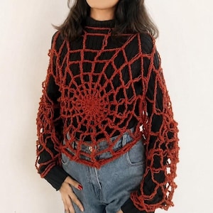 May include: A rust-colored, hand-crocheted spiderweb-patterned overlay worn over a black long-sleeved ribbed mock-neck shirt and blue denim jeans. The open-knit garment features a circular web design that extends across the chest and sleeves.