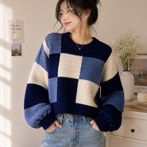 以下が含まれることがあります： ネイビーブルー、ミディアムブルー、クリーム色の大きなチェック柄が特徴の長袖ニットセーター。クルーネック、リブ編みの袖口と裾が施されており、ボタンフライのライトブルーのデニムジーンズと組み合わされています。