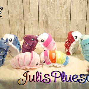 Könnte beinhalten: Eine Sammlung von acht Isopoden-Plüschspielzeugen in verschiedenen Farben, darunter Blau mit gelben Sternen, Lila, Pink, Rot, Hellblau und eine regenbogenfarbene Version. Der Text Julis Pluesch ist unten in einer stilisierten Schrift zu sehen.
