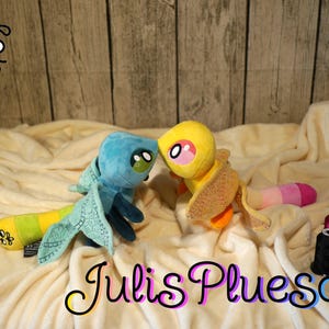 Könnte beinhalten: Zwei Libellen-Plüschtiere, eines blau und eines gelb, mit gemusterten Flügeln und segmentierten Schwänzen. Das blaue Spielzeug hat grüne Augen, das gelbe hat rosa Augen. Der Text Julis Pluesch ist unten sichtbar, dazu Nähgarnrollen und eine Schere.