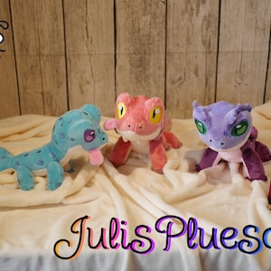 Könnte beinhalten: Drei Plüsch-Echsen in verschiedenen Farben: blau, rosa und lila. Die blaue Echs hat ein gepunktetes Muster. Die rosa Echs hat gelbe Augen. Die lila Echs hat grüne Augen. Die Spielzeuge liegen auf einer weißen Decke mit dem Text "Julis Plusch" in bunten Buchstaben.