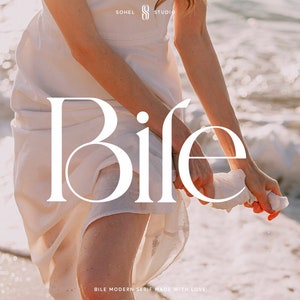 May include: A white dress with the word "Bife" written in a white serif font on the front. The word is centered on the dress and is slightly angled. The dress is being held up by a person's hand. The background is a blurry image of a beach with waves.