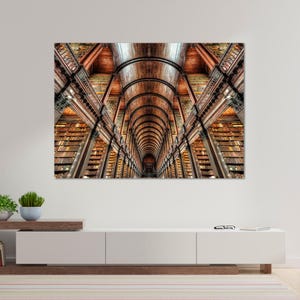May include: A large rectangular wall art print featuring a symmetrical perspective of a long library hall with towering wooden bookshelves, a vaulted wooden ceiling, and rows of books lining the walls in warm brown and golden tones.