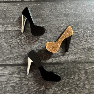 May include: Three high-heeled shoe decorations. Two are black with silver heels, and one is gold glitter with a black heel. The shoes are arranged on a grey wooden surface.