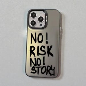 Peut inclure: Coque de téléphone argentée avec bordure noire et texte "NO! RISK NO! STORY" en style manuscrit. Le boîtier possède un module caméra avec trois lentilles et un flash.