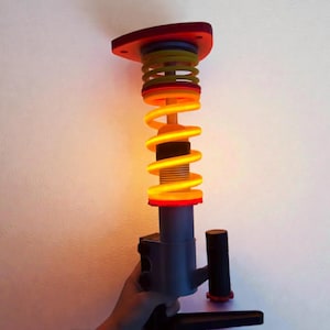 May include: A colorful, illuminated sculpture featuring a spring-like design. The sculpture has a red top and base, with yellow and orange accents. The central element is a glowing, spiraling light, creating a vibrant and eye-catching display.