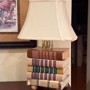 May include: A unique lamp with a cream-colored lampshade, resting on a base made of stacked vintage books. The books are various colors, including brown, yellow, and purple. The lamp has a brass-colored neck and a wooden base.