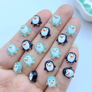 May include: A selection of small penguin charms in black and light blue. Each charm has an orange beak and white belly. The charms are arranged on a light background. The penguin charms are suitable for crafting.