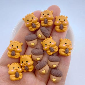 May include: A selection of small, orange squirrel-shaped embellishments, each holding a brown nut. There are also brown acorn-shaped embellishments with yellow heart details. These decorative pieces are suitable for crafting and scrapbooking projects.