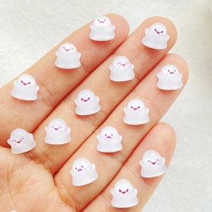 May include: Small, translucent white ghost-shaped charms with pink smiling or frowning faces. The charms are displayed on a light surface, possibly for crafting or decoration. Each charm is a unique, miniature design.