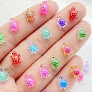May include: A selection of tiny, brightly coloured turtle charms. The charms are translucent and available in a range of colours, such as pink, orange, red, green, blue, and purple. The turtle shells feature a patterned design. These charms are likely for crafts or jewellery.