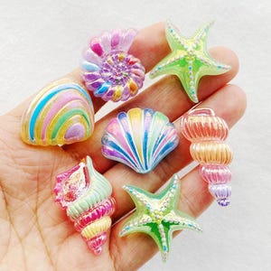 Puede incluir: Una colección de adornos coloridos e iridiscentes de conchas marinas y estrellas de mar. Las conchas presentan una variedad de diseños, incluyendo una espiral, una forma de abanico y una caracola. Las estrellas de mar son verdes con detalles amarillos.